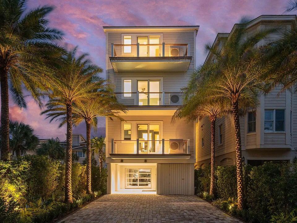 Exterior of multi-story house with driveway lined with palm trees at night.