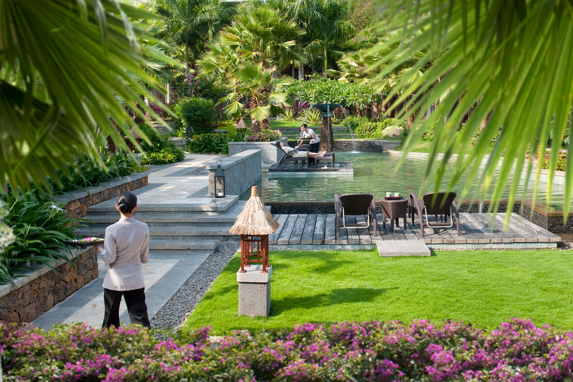 An employee with black hair in a bun carries a tray through the gray path in the lush SPA Garden, towards the pool where a guest reclines.