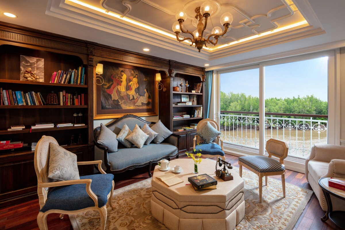The library on a Uniworld cruise has dark wood bookshelves set into a wall. Antique chairs and couches provide seating next to large windows.