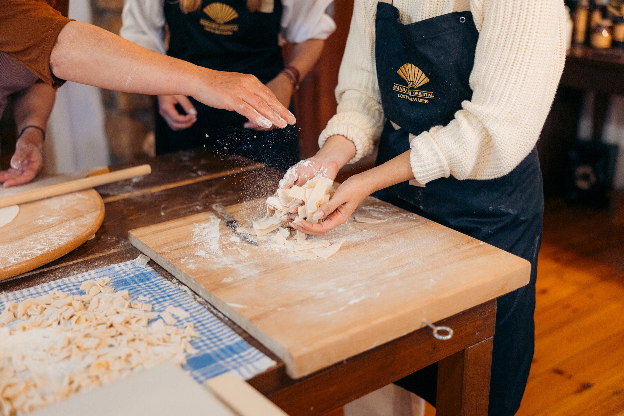 Shoulder-down view of a cooking class as a woman in a white sweater and a Mandarin Oriental apron handles pasta over a floury cutting board.