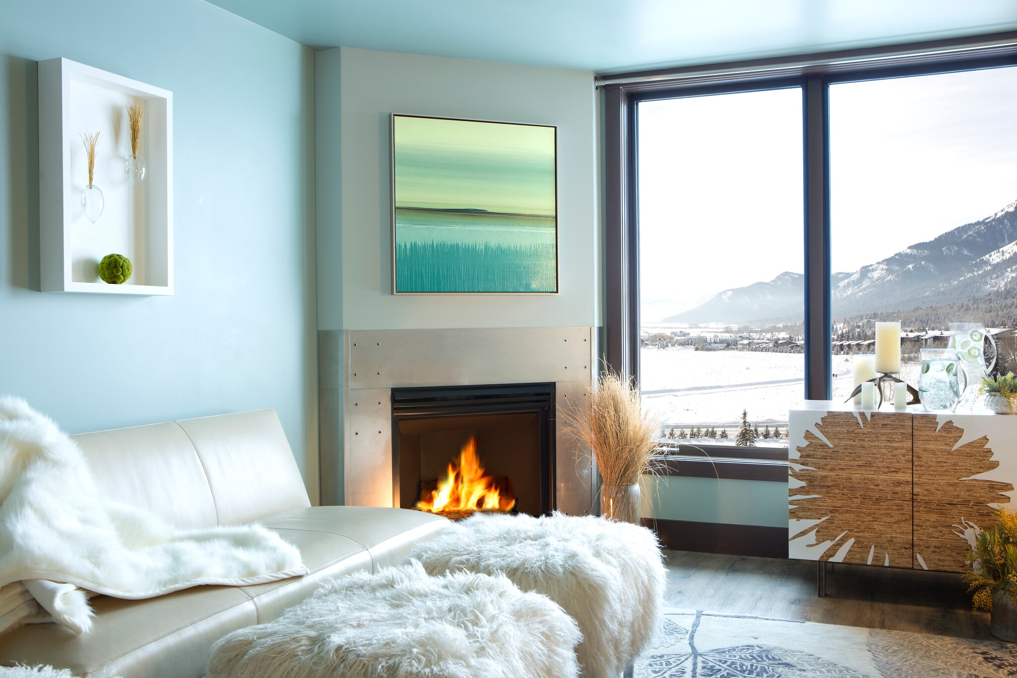 The living area of a guestroom has pale blue walls and white furniture, including fuzzy ottomans. Snowy mountains peek in the window.