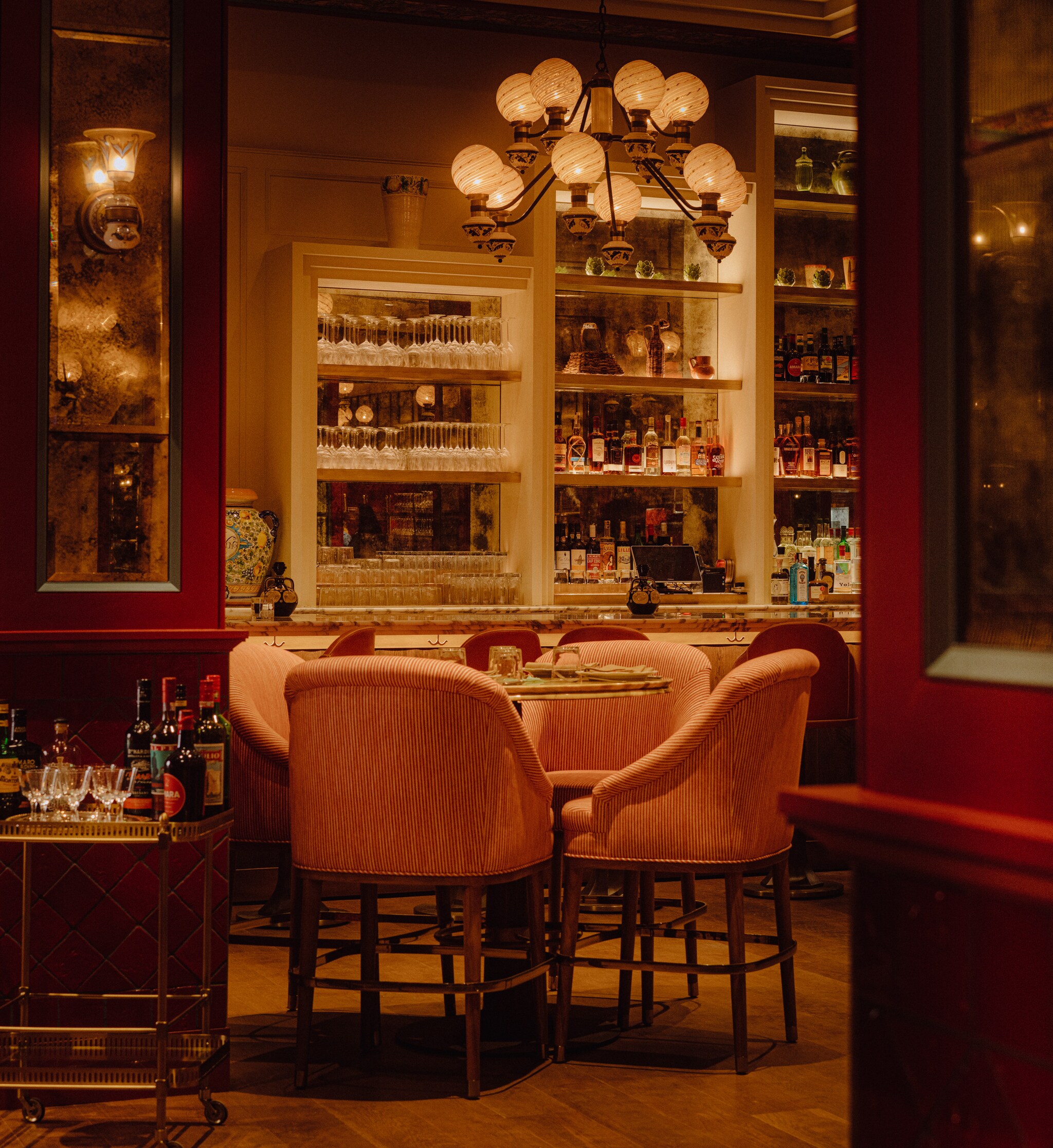 In dim orange light, pink armchairs crowd around a circular table. Behind, a bar is outfitted with bottles of spirits and clear glasses.