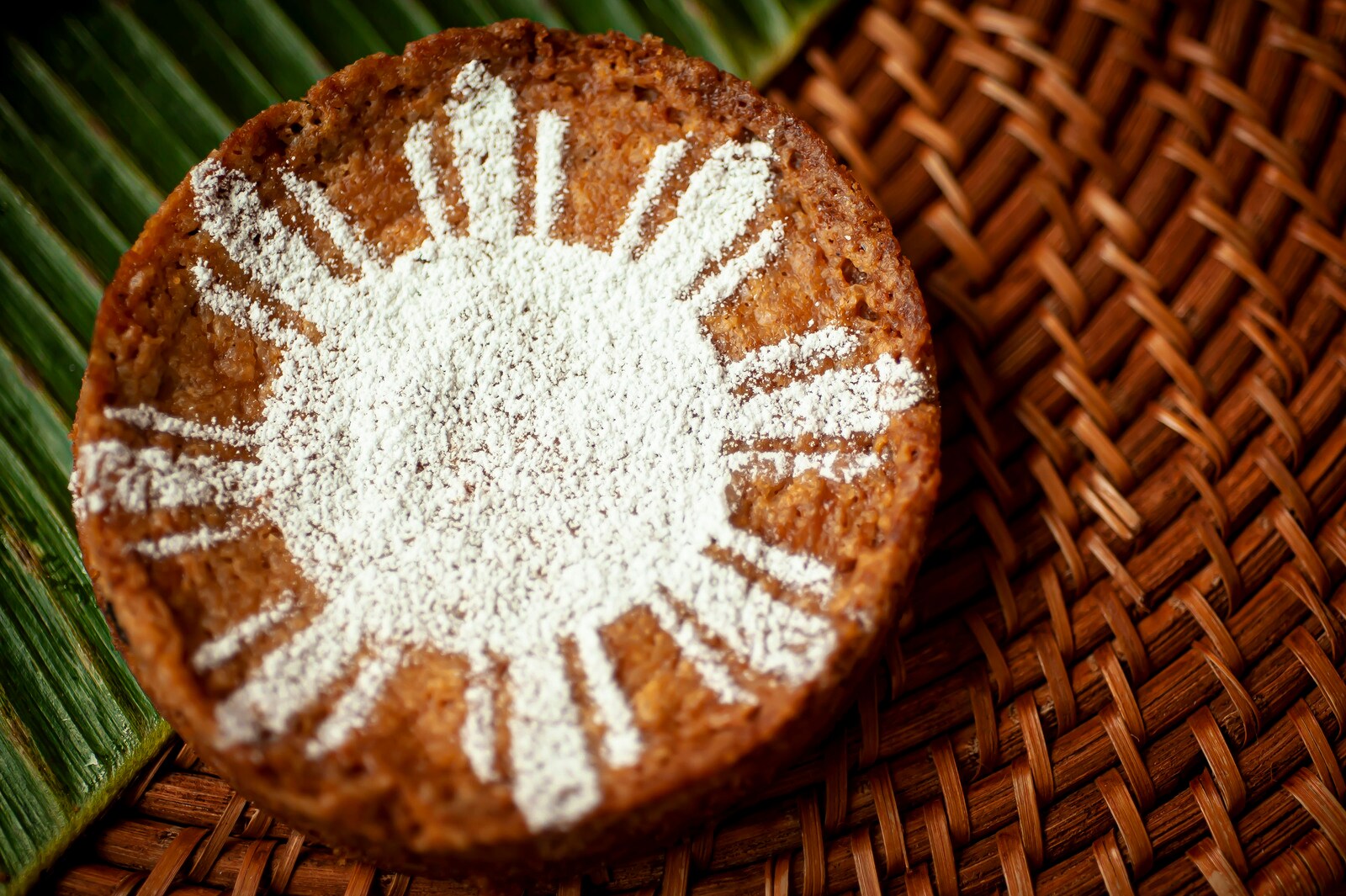 Powdered sugar forms a sun on a small, circular cake baked to a deep golden brown and arrayed on a leaf and woven placemat. 