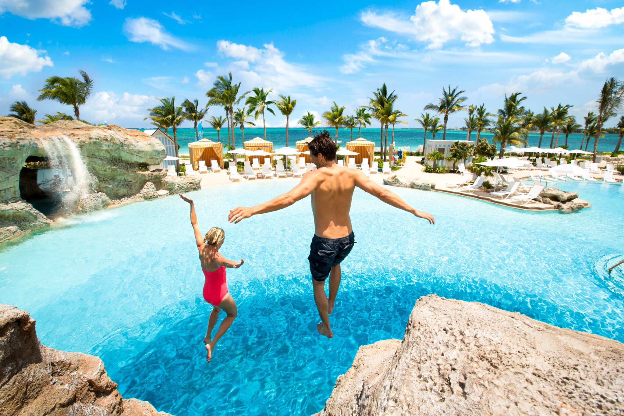 View from behind as a young white father and his daughter jump off of a rocky ledge into an outdoor pool. The ocean is visible beyond.
