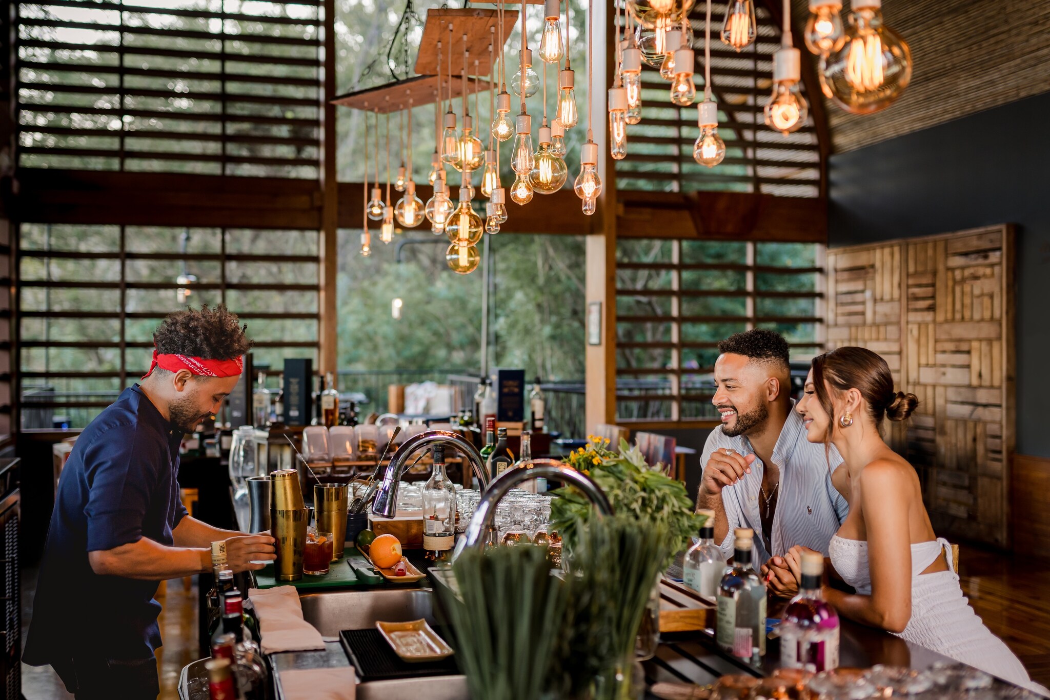 At a trendy hotel bar, a bartender mixes a cocktail for a Black man and a Latina woman, fashionably dressed and leaning close.
