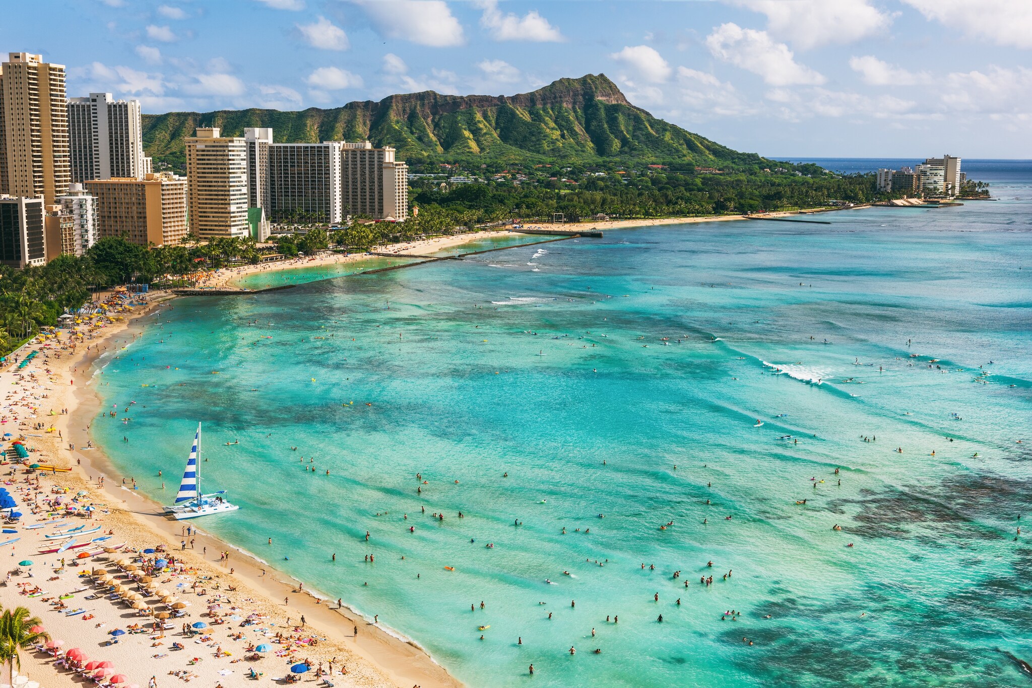 View over the beach at Honolulu with Diamond Head on the horizon. A small sailboat is about to take off and hundreds of beachgoers play.
