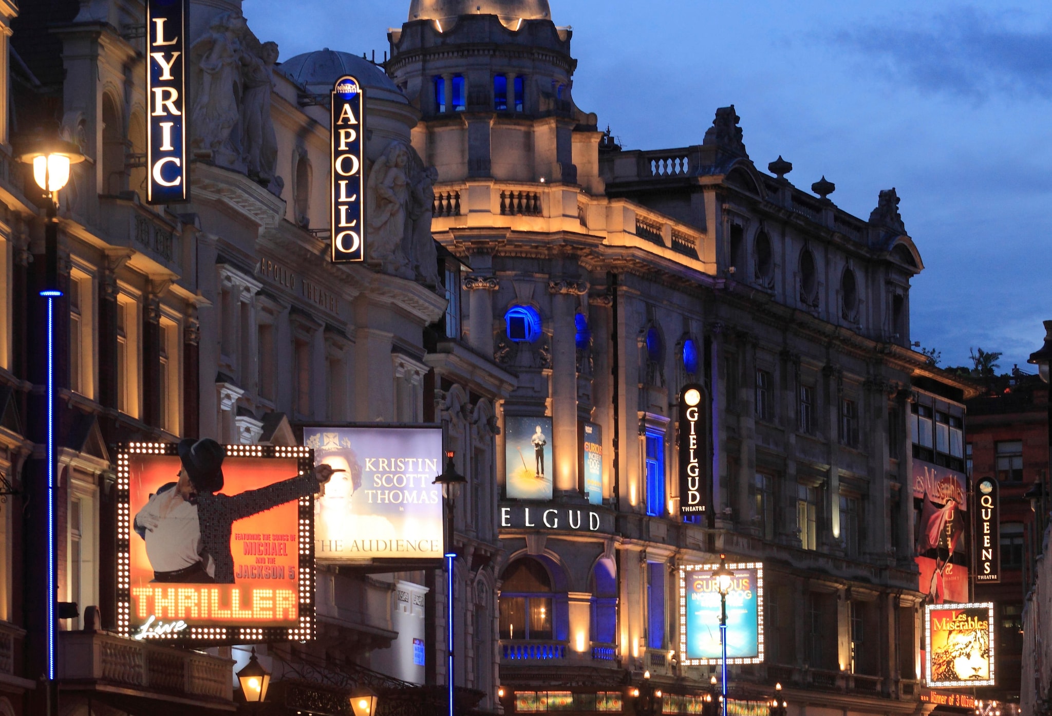 View down a West End street in the evening, with glittering marquees for theaters and musical productions lit up in the gloom.