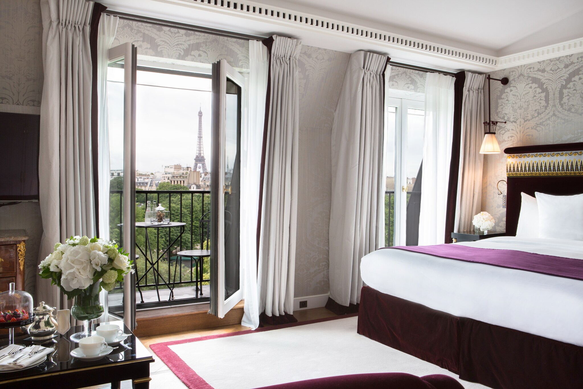 A premier junior suite is decorated with ornate gray wallpaper and purple accents on white bedding. Balcony doors overlook the Eiffel Tower.