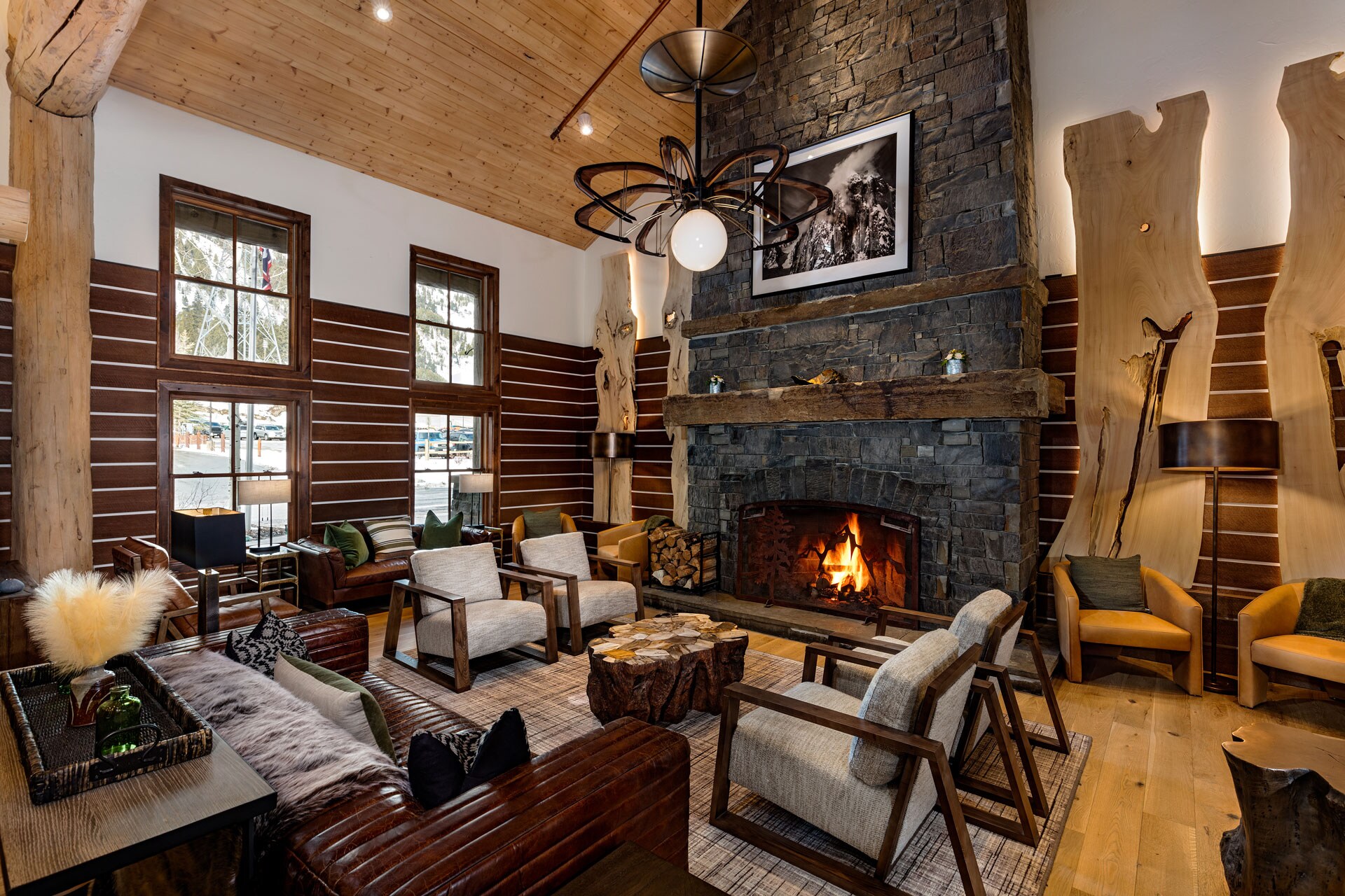 In a high-ceilinged lounge space, gray armchairs and a brown leather couch surround a stump table and a roaring stone fireplace.