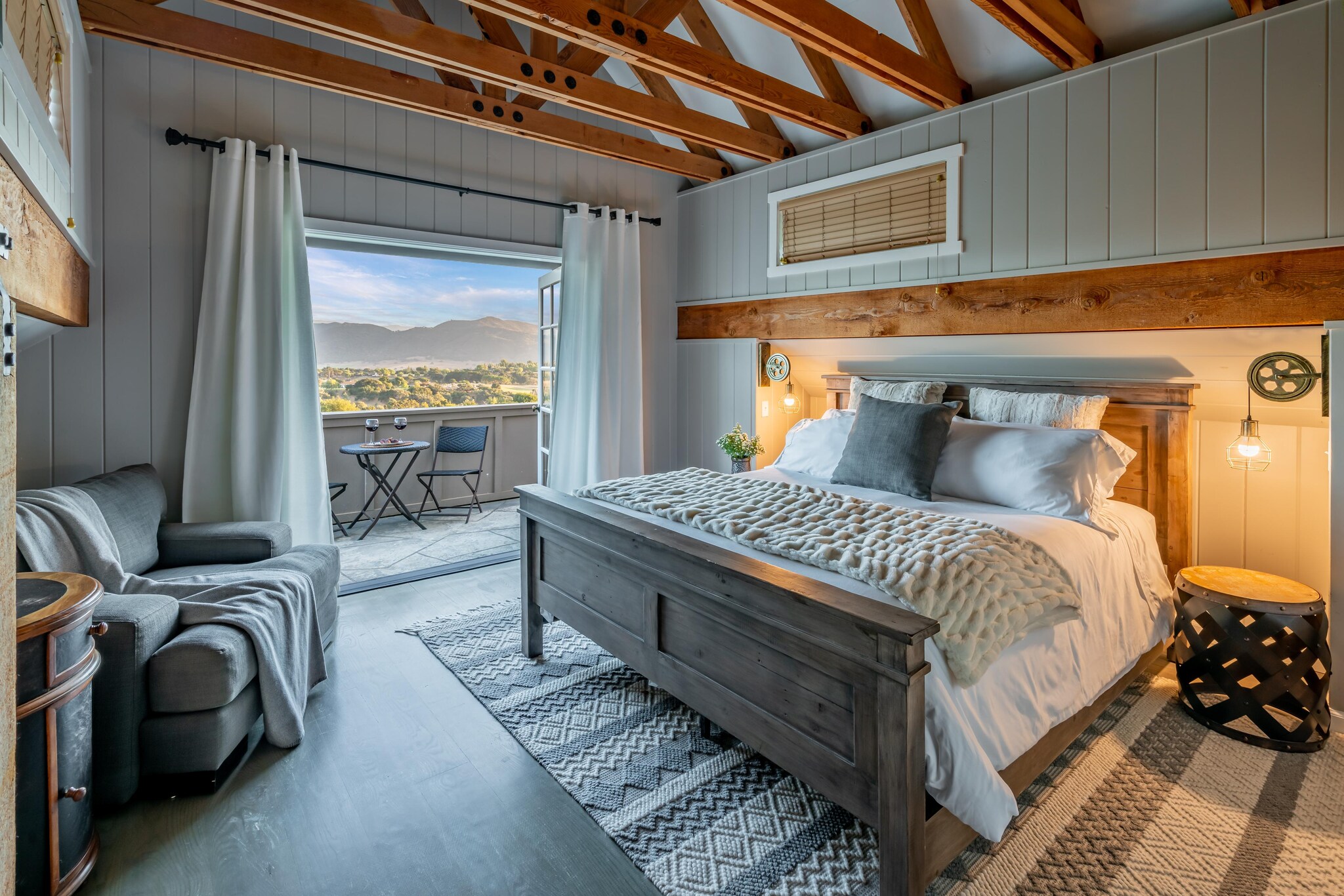 Cottage style rustic bedroom with private balcony featuring a bistro table and two chairs with view of the surrounding countryside and mountains.