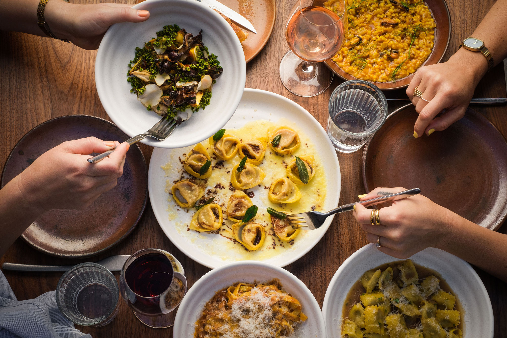 Overhead view of a table covered with drinks and dishes of pasta. Hands reach across with forks to try salad and tortellini.