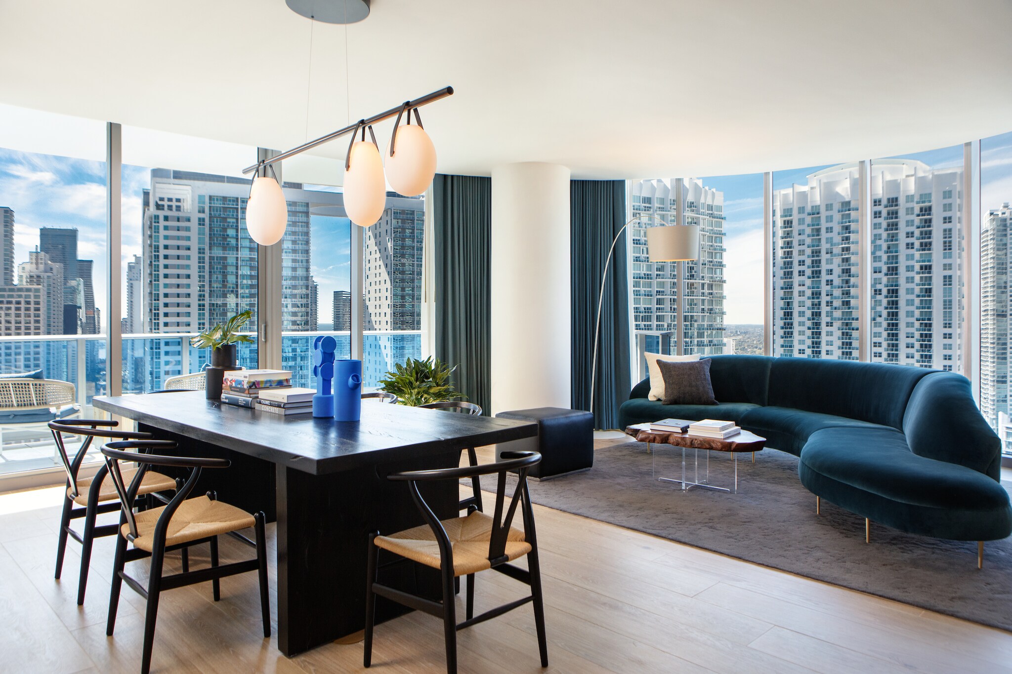 The airy Penthouse Suite living room is arrayed with a large dining table, a large curved blue velvet couch, and floor-to-ceiling windows.