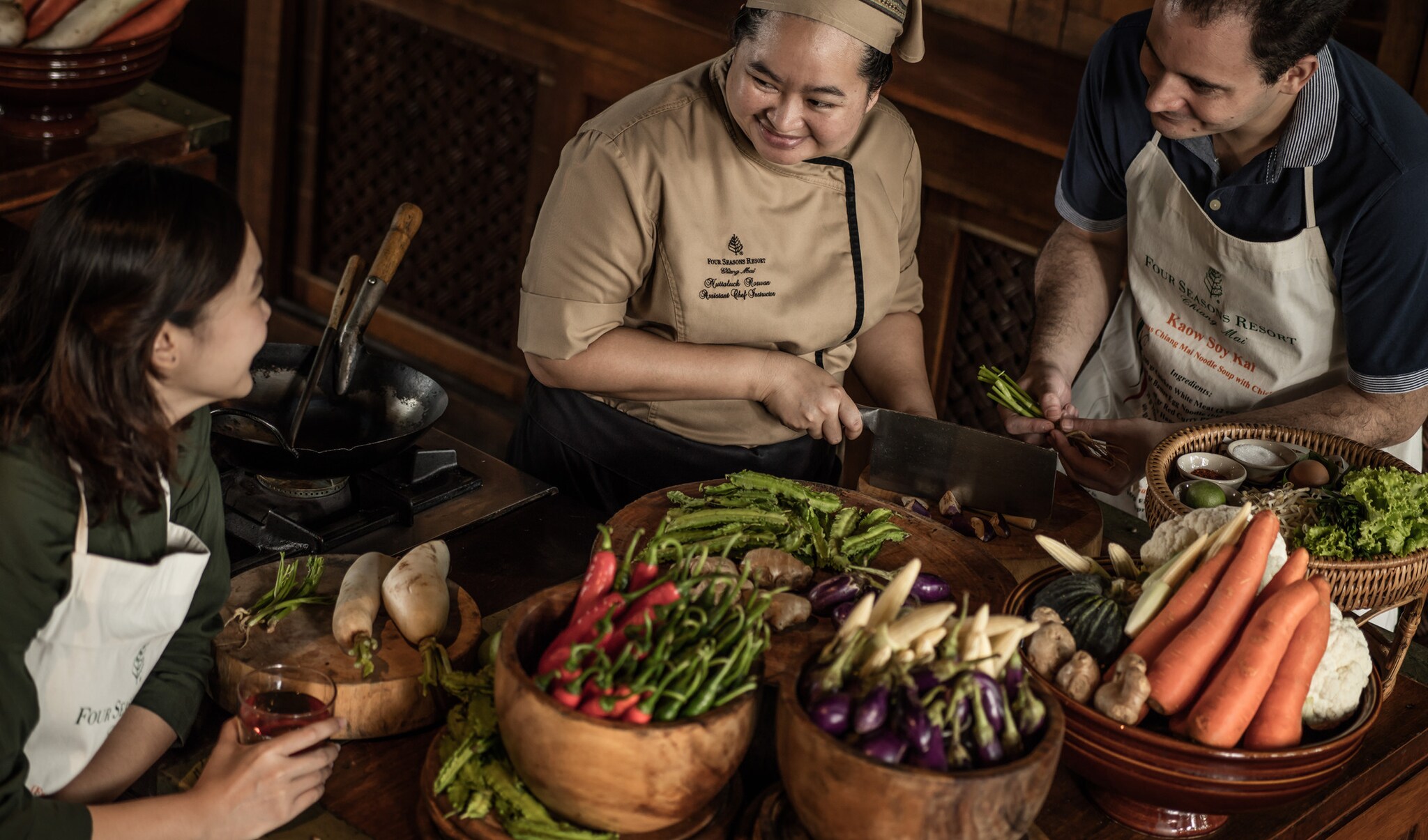 Amidst wooden bowls of fresh produce and spices, an Asian woman in a brown chef’s uniform leads guests in aprons through a cooking class.