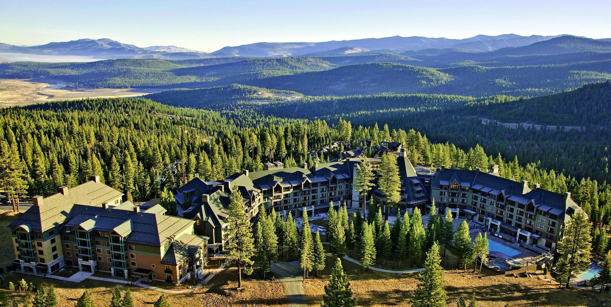 Aerial view of the hotel, nestled in forest between mountains. The buildings are joined to form a long curve around pools.