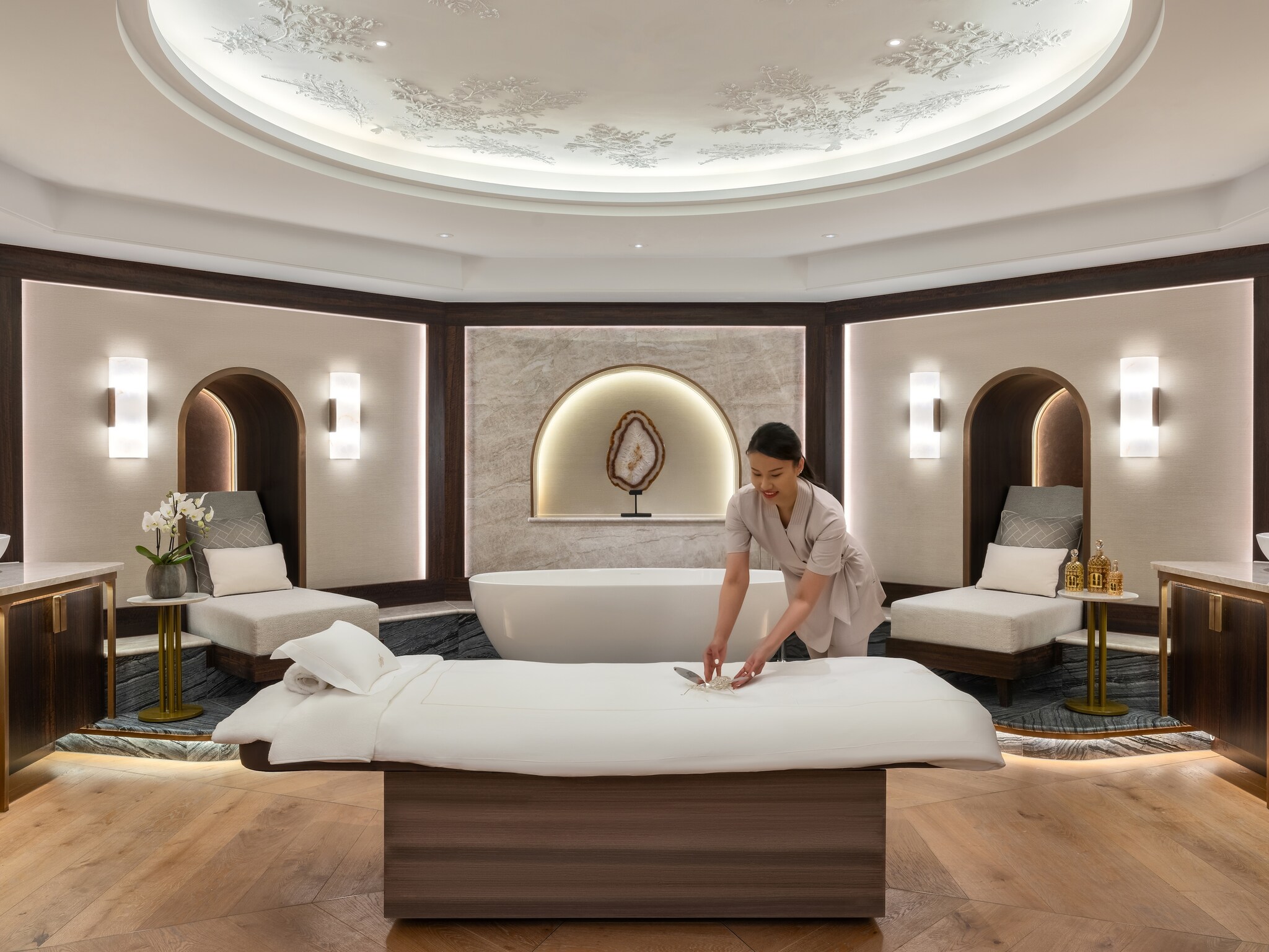 In the spa, a staff member in a gray uniform arranges an ornamental dragonfly on an elevated bed.