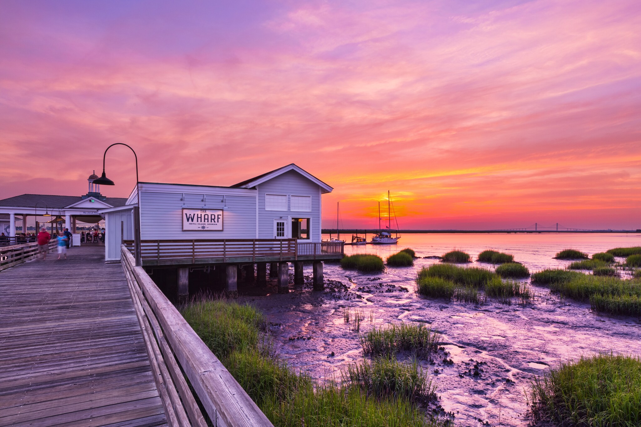At sunset, The Wharf waterfront restaurant is a small blue building set on a pier over reeds and orange-tinged water.