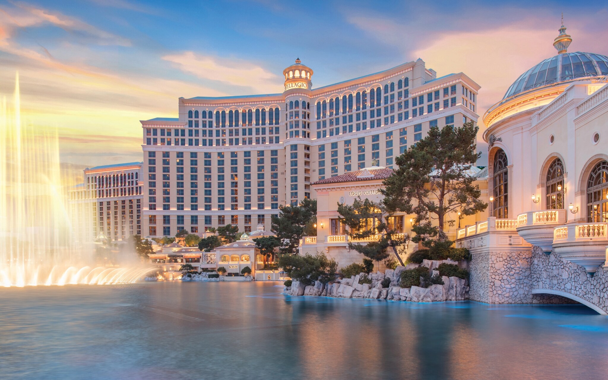 An external view of the Bellagio at sunset looks across a pond to fountains splashing in front of the grand main building.