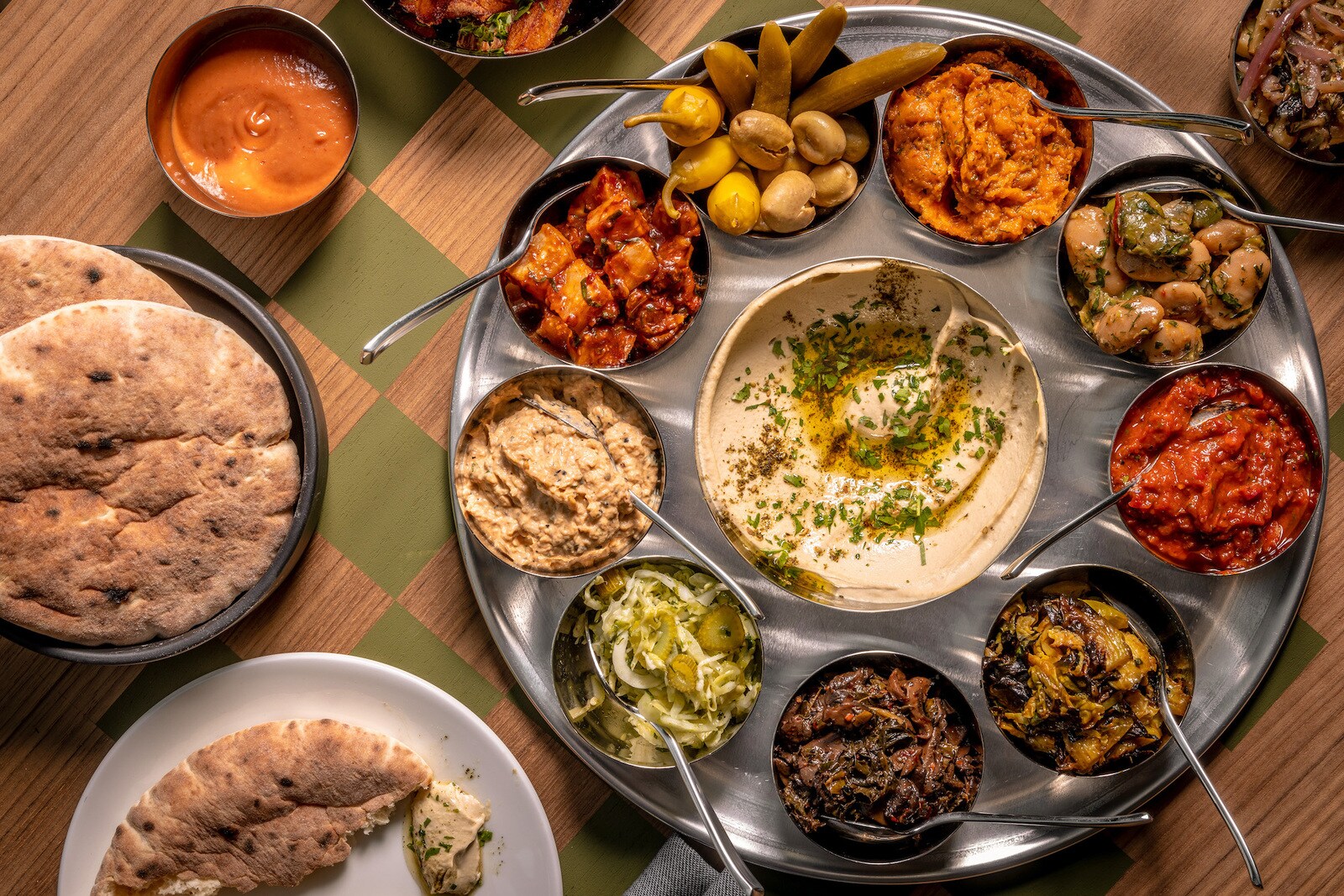 Overhead view of a round silver tray of hummus, olives, beans, and other dips to accompany nearby plates of pita.