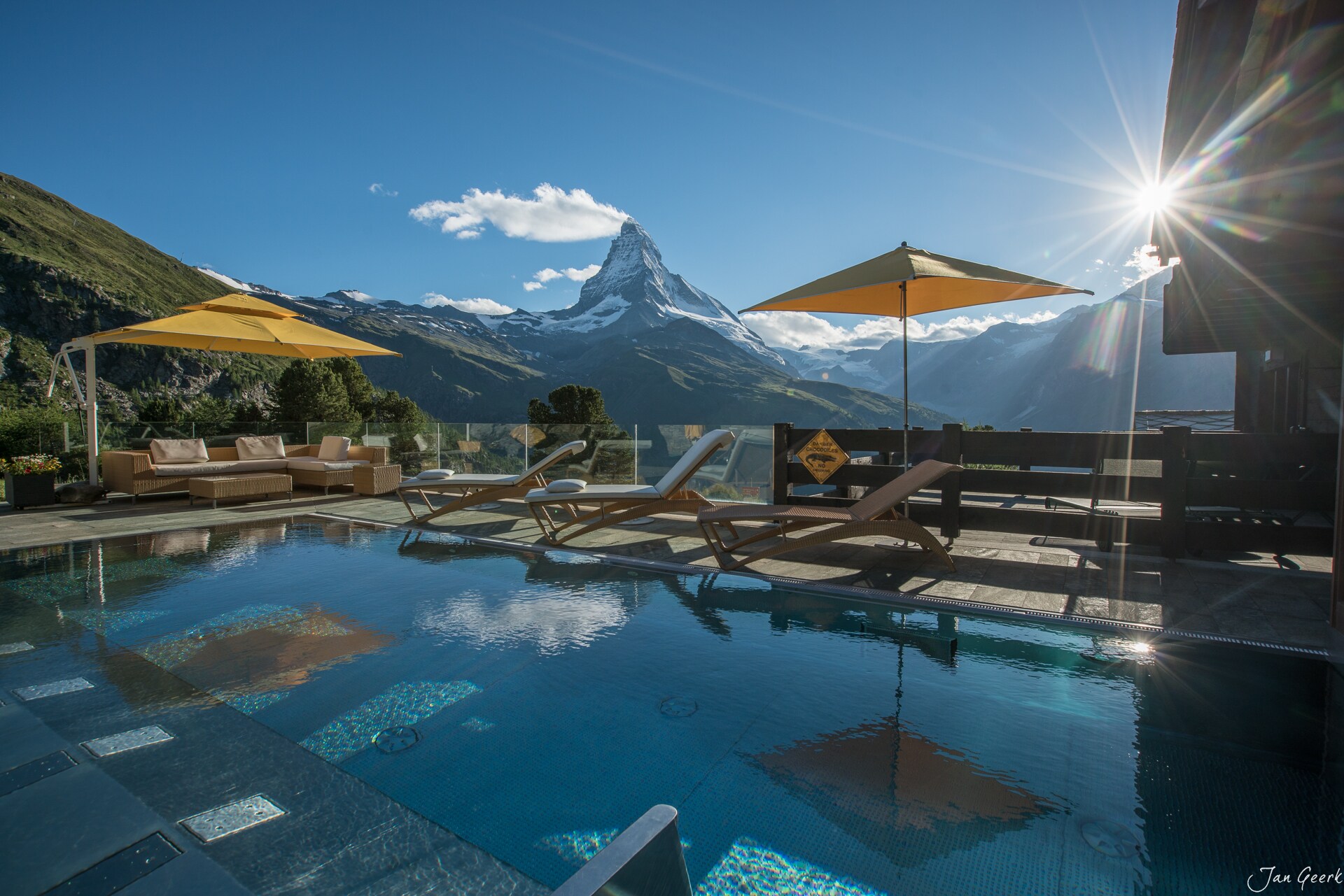 A heated outdoor pool is slightly shaded by square yellow umbrellas. In the background, green mountainsides rise occasionally to snowy caps.