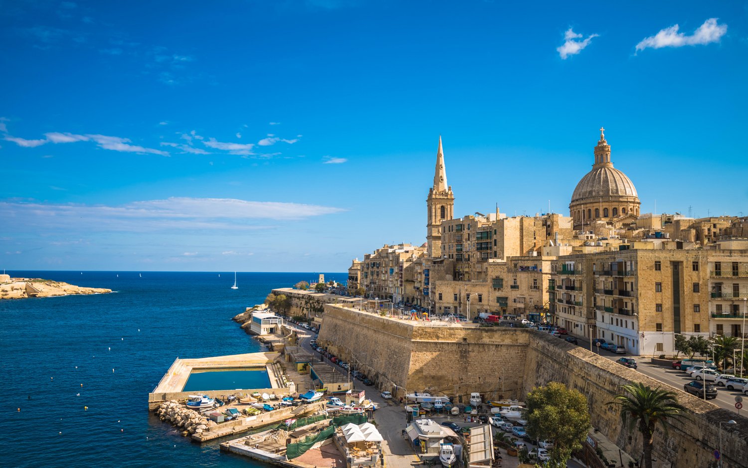View of the sandy-brown Basilica of Our Lady of Mount Carmel and matching nearby walls bordering the bright blue Mediterranean water.