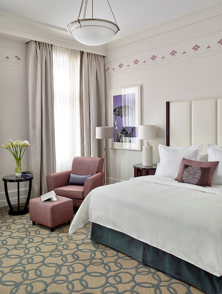 A guest room is decorated in white and gray, accented with pink cushions and seats and a vase of calla lilies on a small end table.
