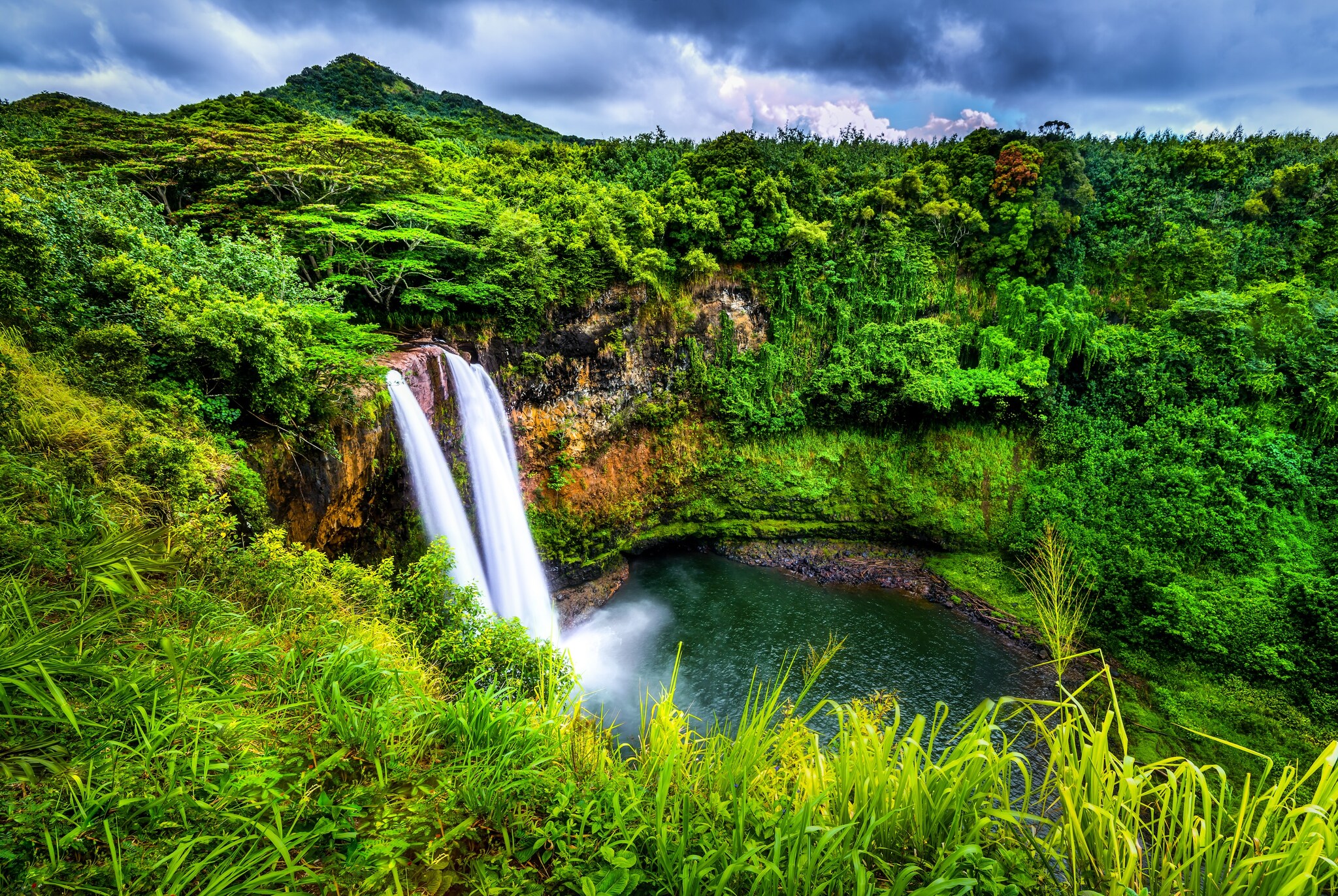 Surrounded by bright, dense forest greenery, two streams of Wailua Falls pour off a cliff into a round, blue-green pool.