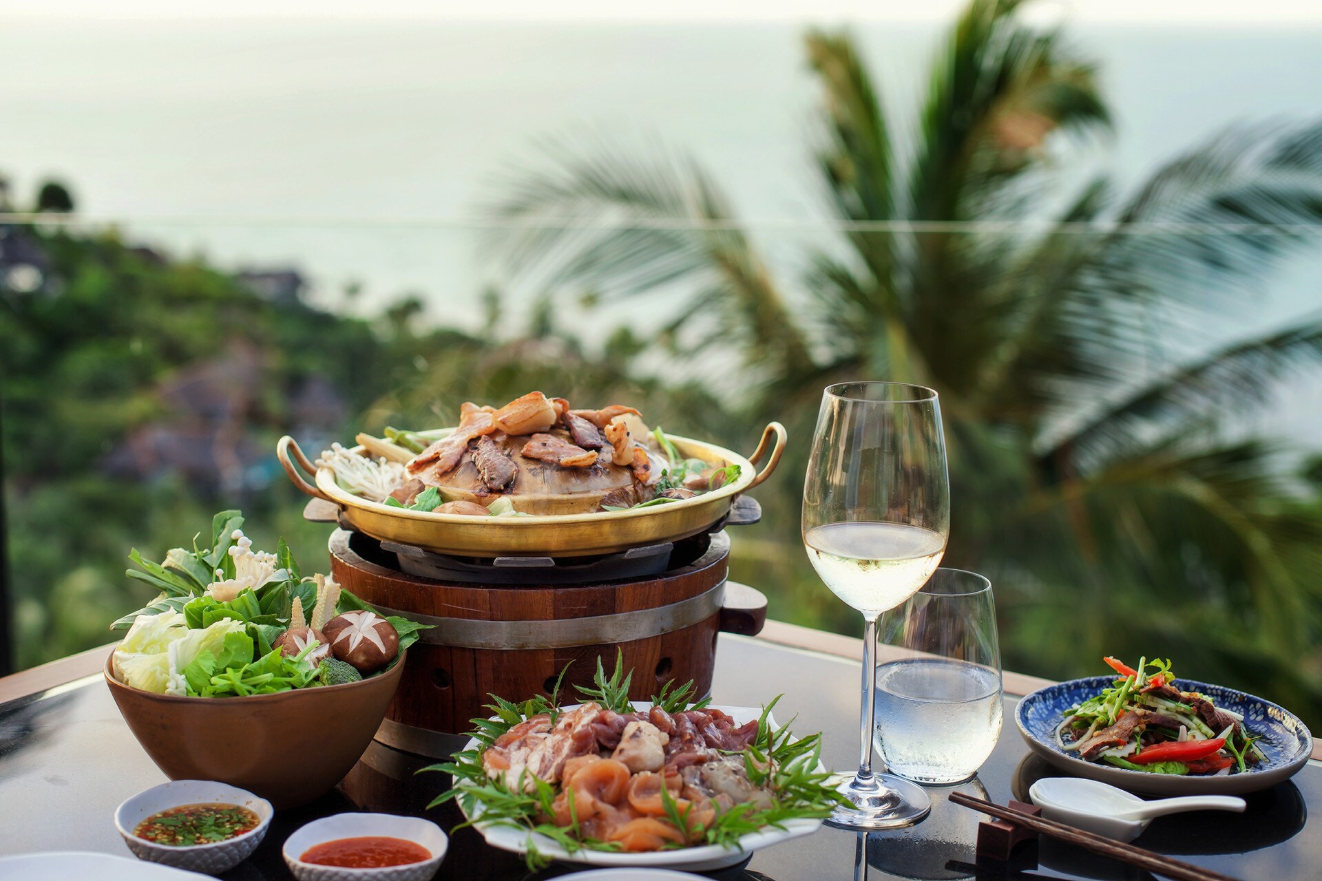 Close-up of an outdoor table set with sprouts, vegetables, and delicately cooked meat, as well as a glass of water and a glass of white wine.