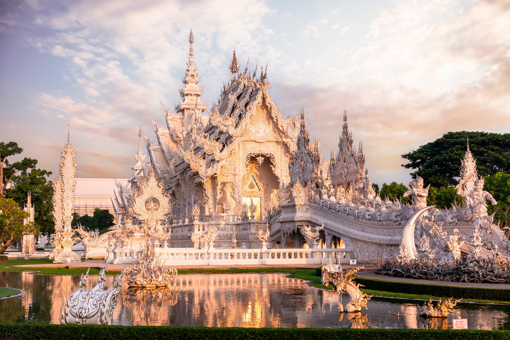 Wat Rong Khun is a white Buddhist temple with incredibly detailed carvings, surrounded by a glassy pool reflecting a sunset.
