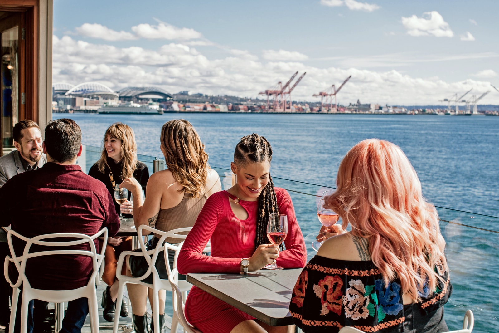 A Black woman with long braids and a white woman with long pink hair sip rosé at a table overlooking the water.