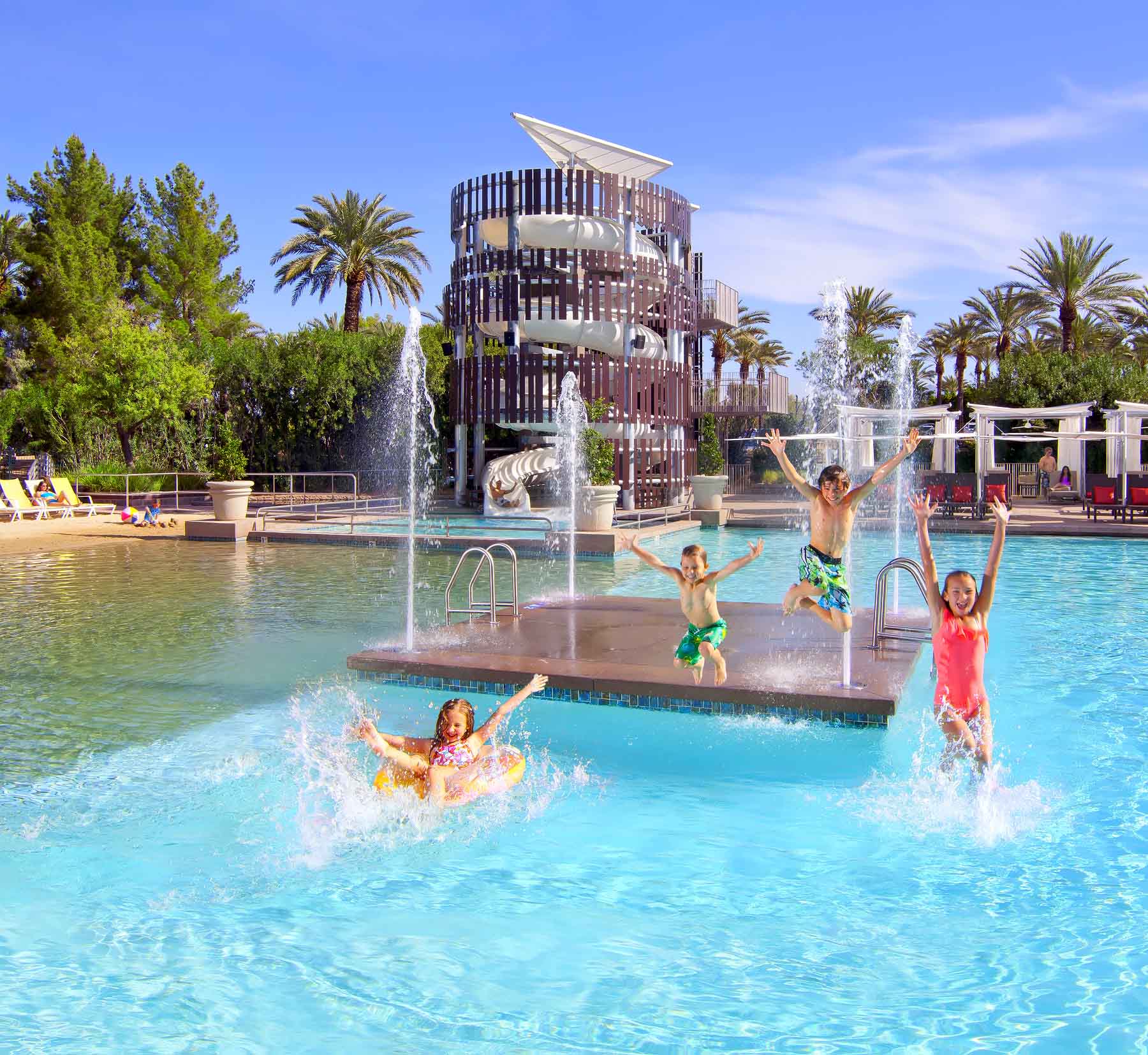 Four white children, two girls and two boys, all wearing brightly colored swimsuits, jump off a fountain platform into an outdoor pool.