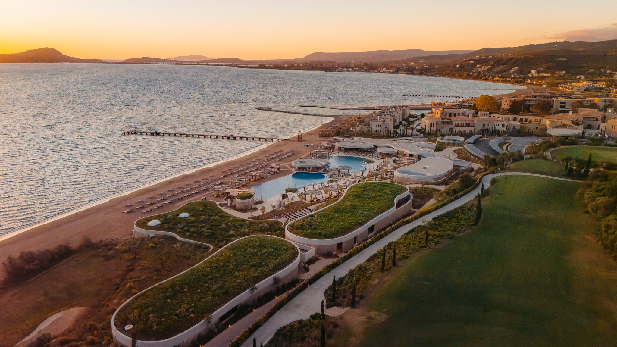Aerial view of the Mandarin Oriental, Costa Navarino at sunset. Grass grows on the roofs of ovular buildings, and pools overlook the beach.