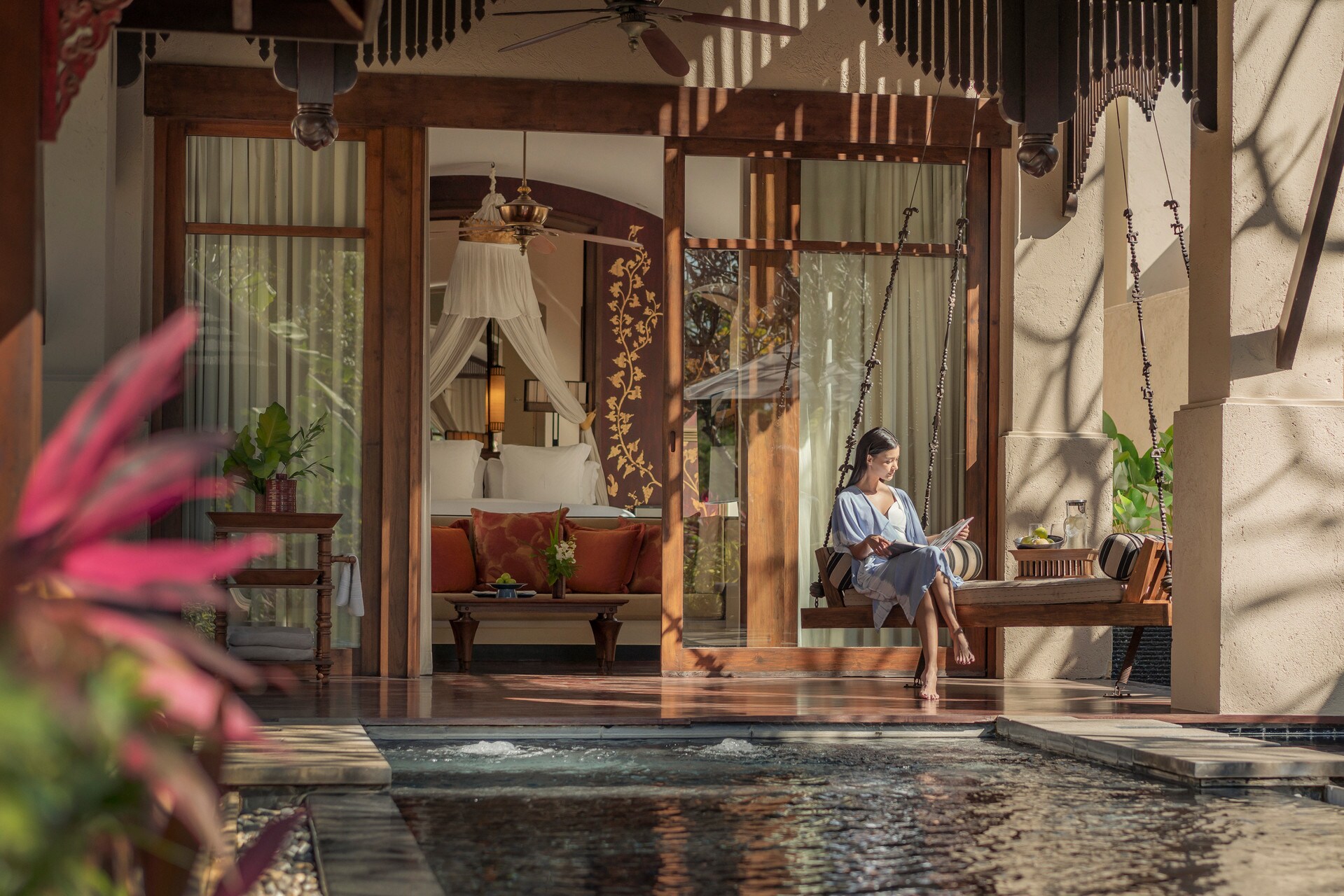A woman with long, dark hair sits on a covered verandah on a hanging bench, reading next to a pool set into the ground.
