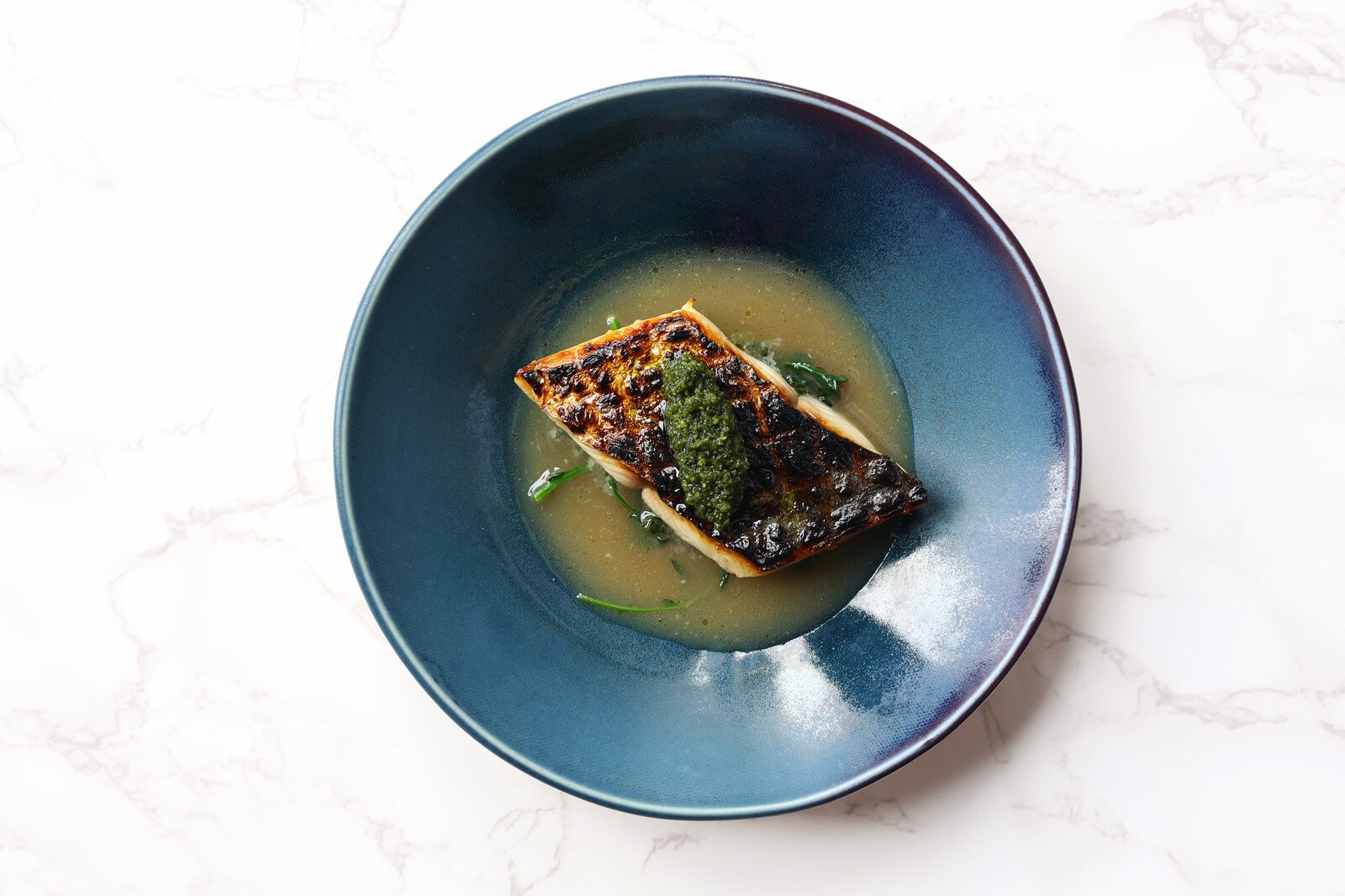 Overhead view of a rhomboid filet of mackerel, finely charred in a grid, sitting in a light yellow broth, and topped with a green paste.