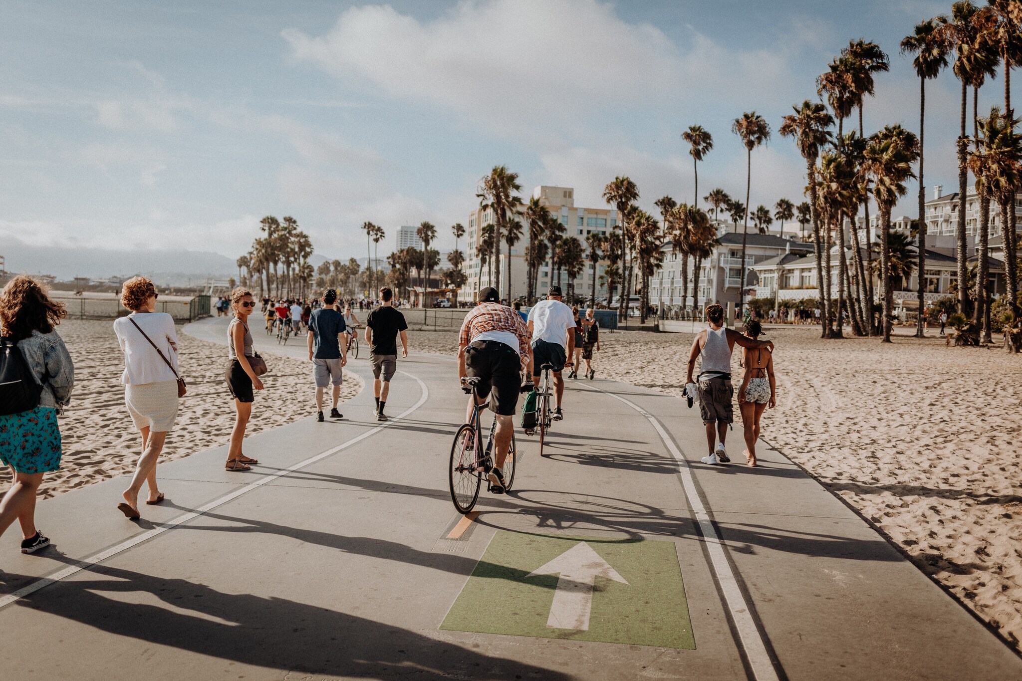 View from behind as bicyclists and walkers travel away from the viewer along the boardwalk between stretches of sand and palm trees.