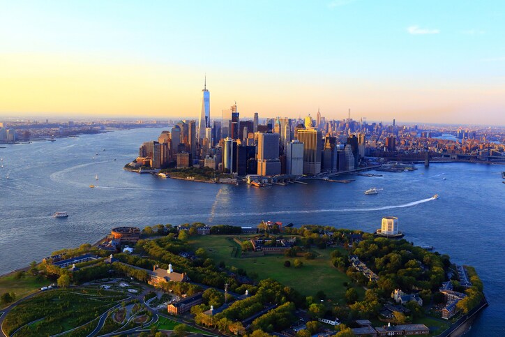 At sunset, the sky is an ombre of colors and the water of New York Harbor is bright blue around the grass and trees of Governor’s Island.