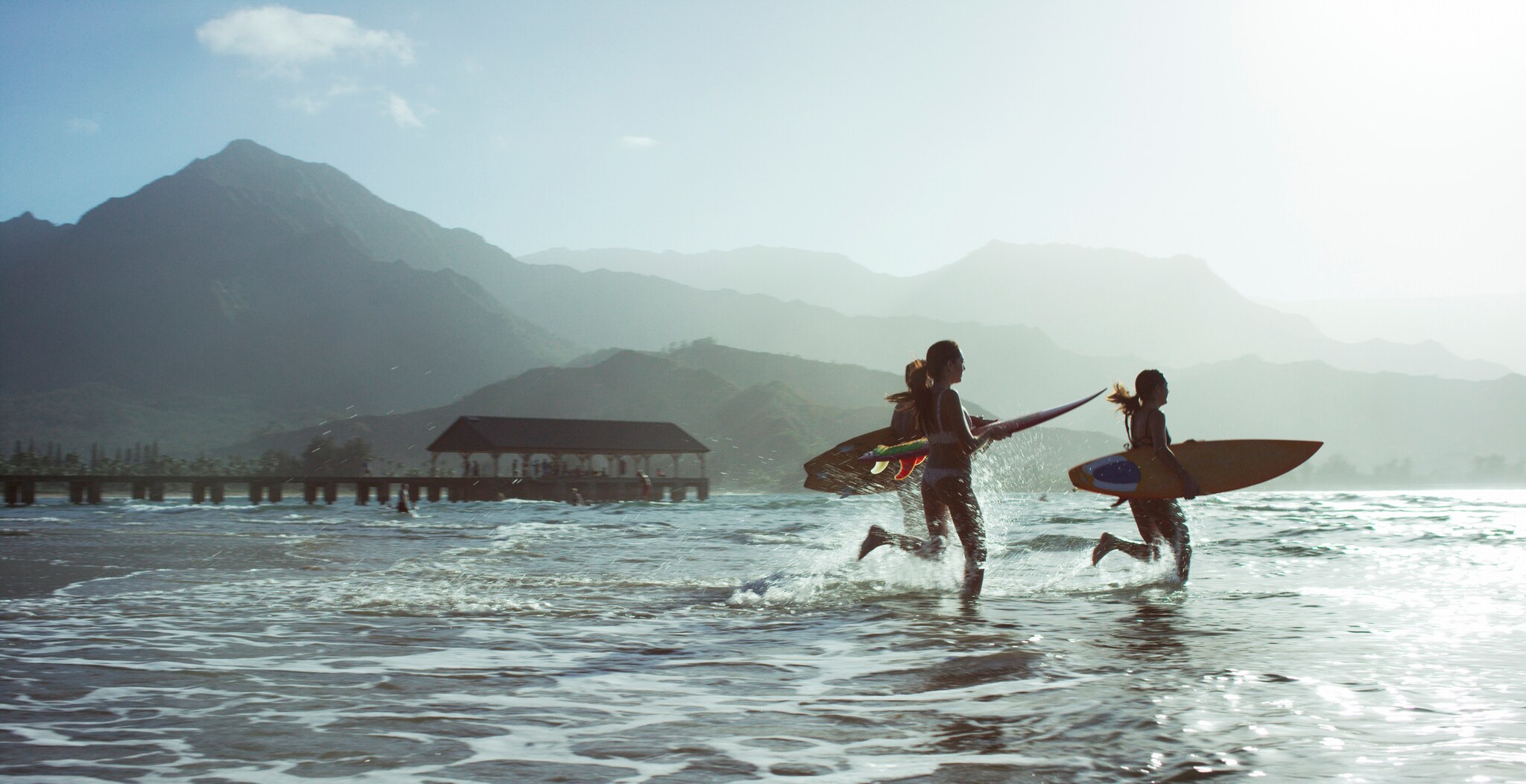 Three children are silhouetted against misty morning light as they run with their surfboards into the ocean.