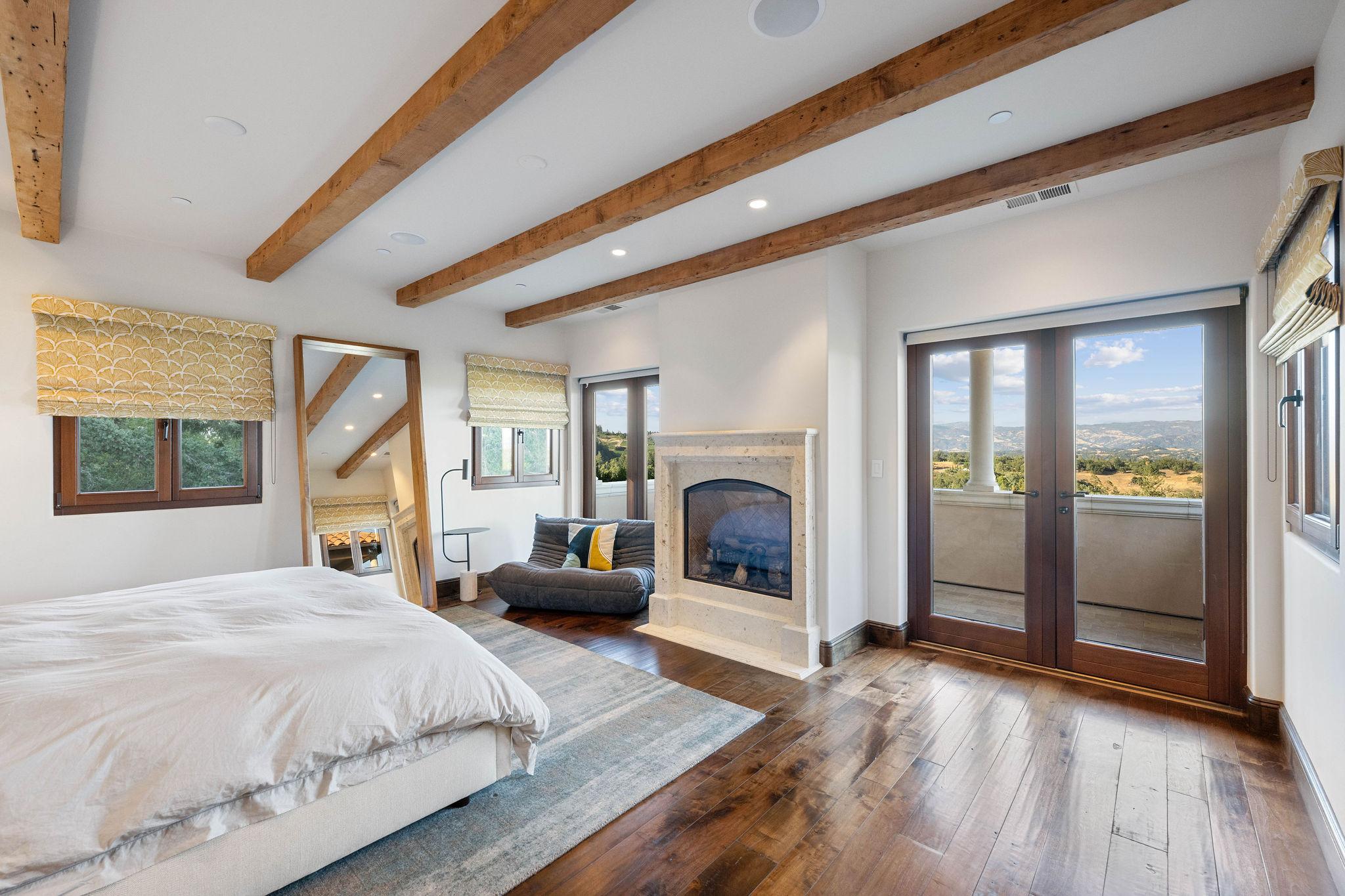 Interior of rustic bedroom with open beams, fireplace, and French doors leading to outdoor balcony.
