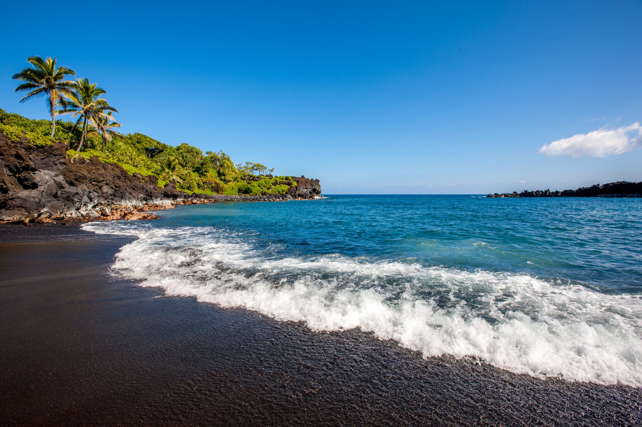 Surf crashes onto the dark sand of a beach. Nearby, a rocky, densely forested hillside curves out towards the horizon.