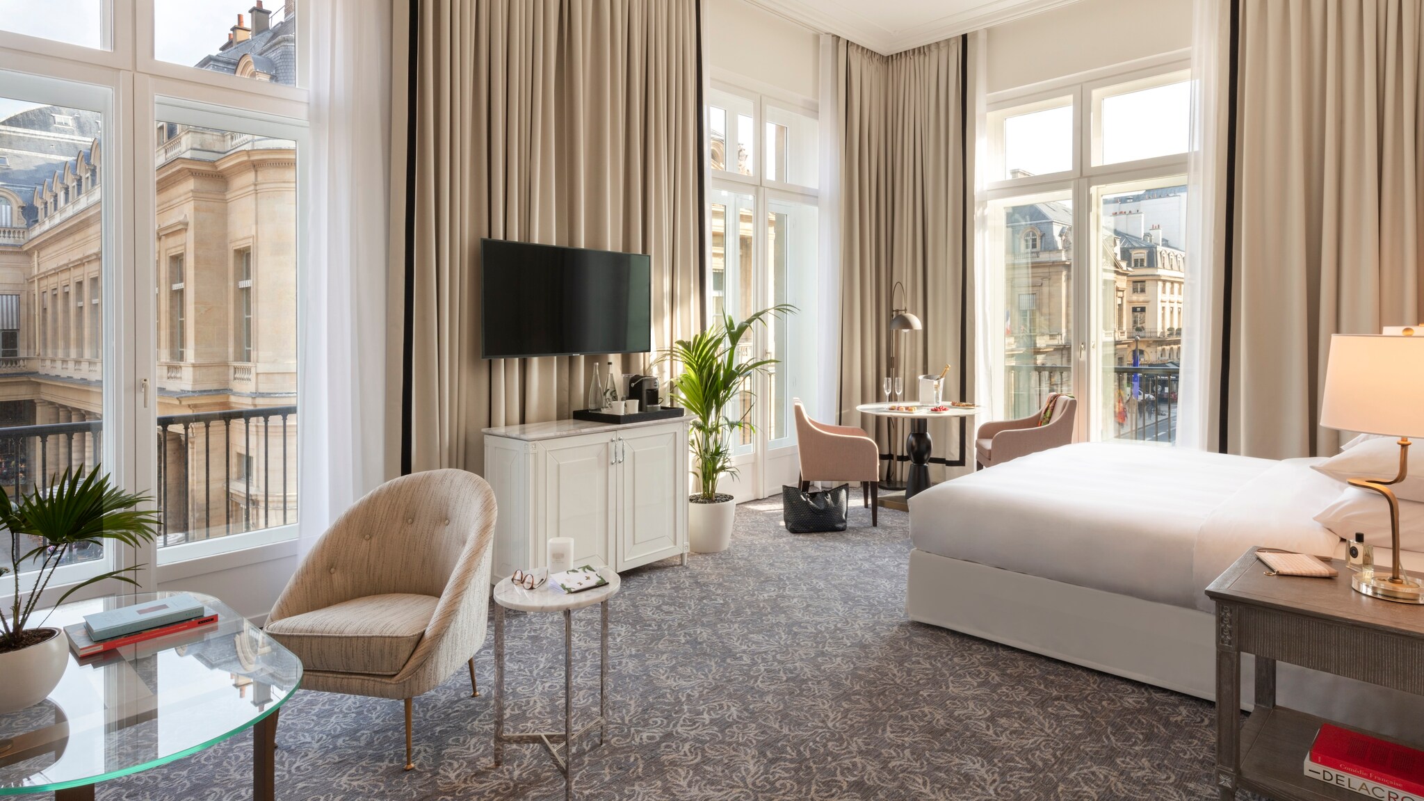 In the Palais Royal Suite, thick white curtains drape around floor-to-ceiling glass doors opening onto balconies. A large bed faces a TV.