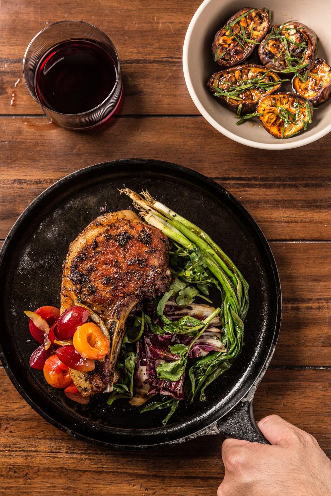 A thick, bone-in cut of meat rests on a cast-iron pan, flanked by small red and yellow tomatoes, broccolini, and radicchio.
