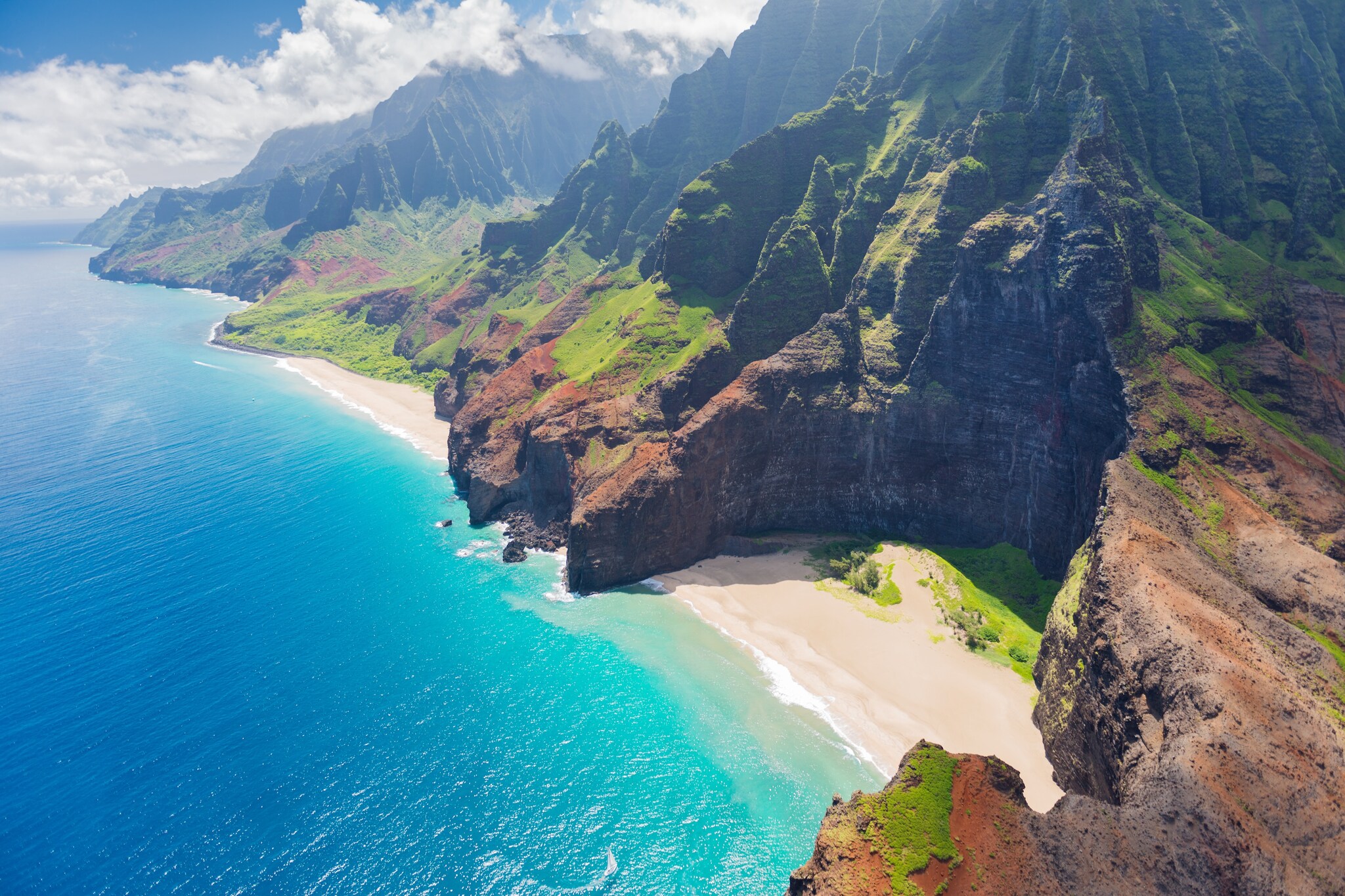 Sunny aerial view of Nā Pali Coast State Wilderness Park, which includes a half-circle of steep cliffs around a beach.