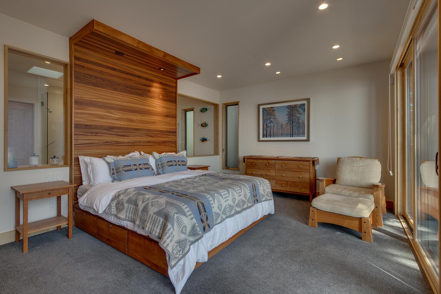 A bedroom has a large bed with a ceiling-height wooden headboard. An armchair and ottoman sit next to sliding glass doors.