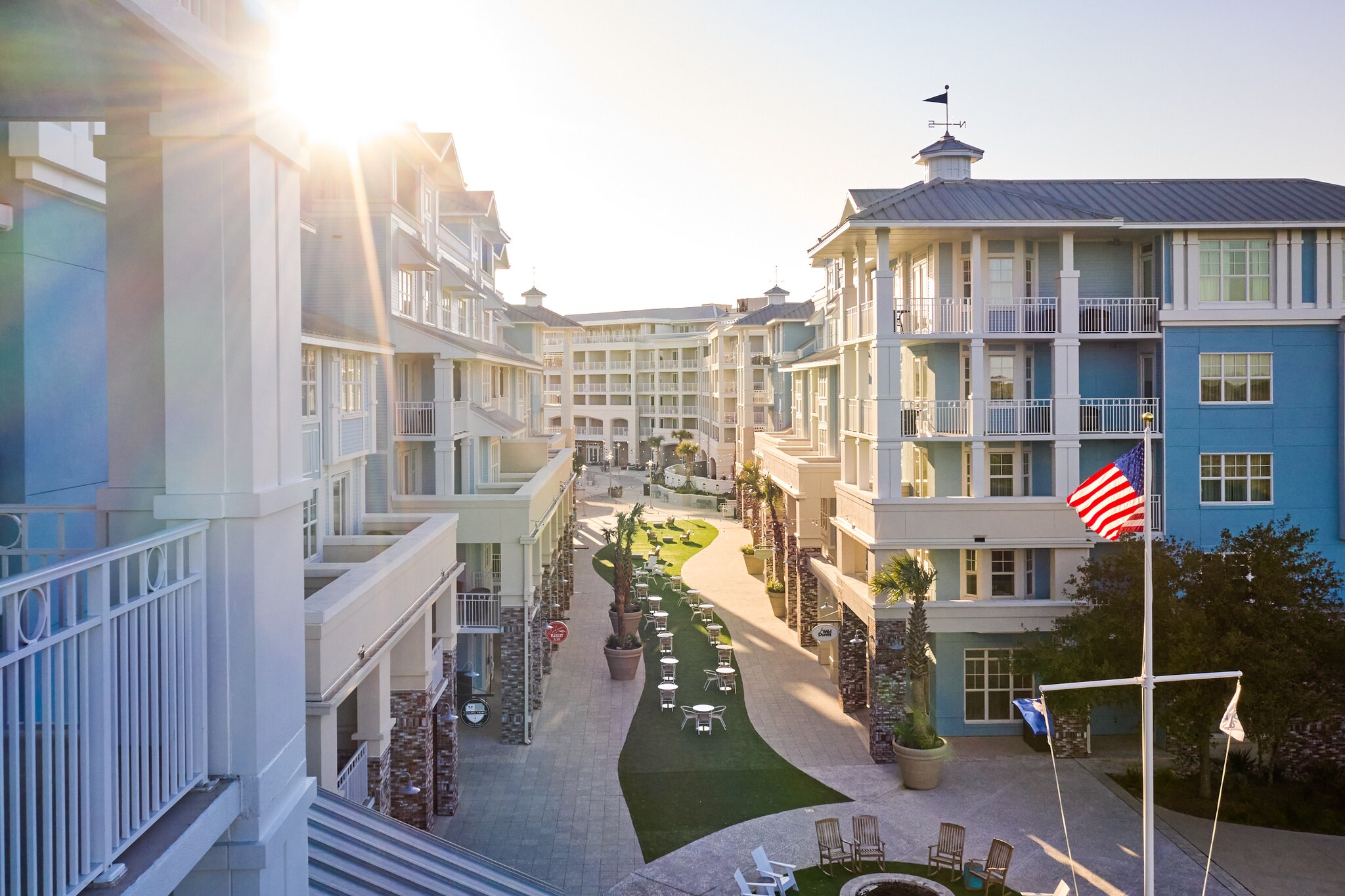 Exterior of Wild Dunes Resort. Sunlight streams through a channel between buildings, onto a wavy strip of grassy and small white tables.