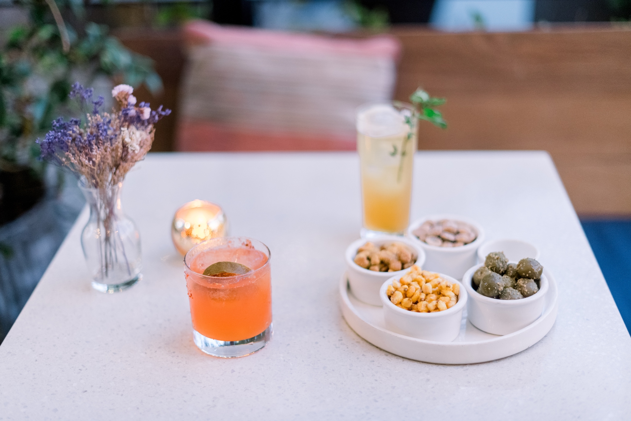 A square white table is arrayed with a small vase of dried flowers, orange and yellow cocktails, and a tray with ramekins of nuts and olives.