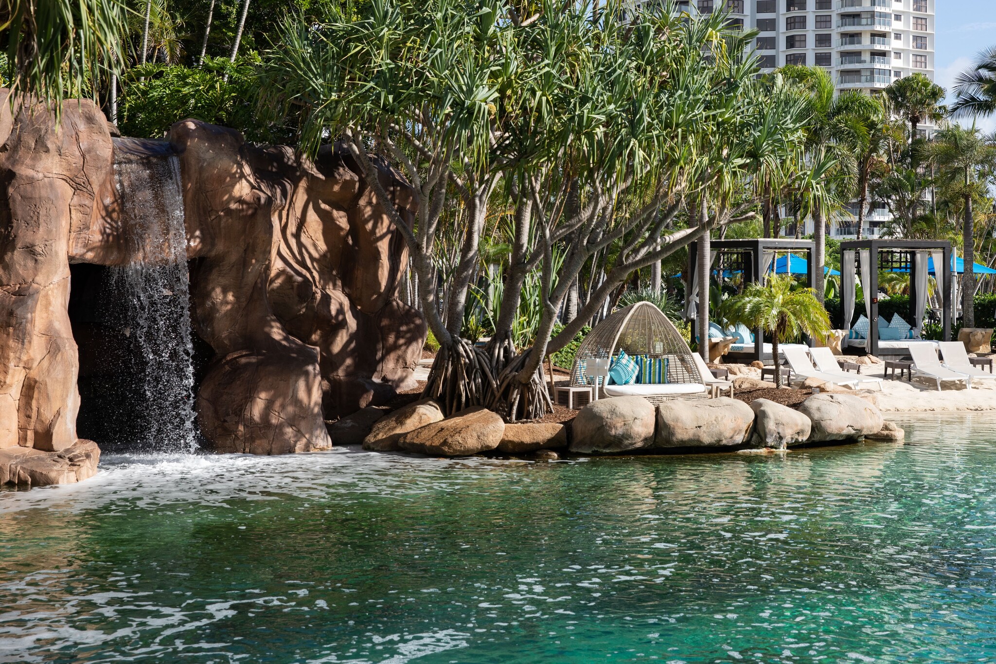 The lagoon pool features a waterfall cascading out from a densely planted area atop a red-brown stone structure. Cabanas are arrayed nearby.