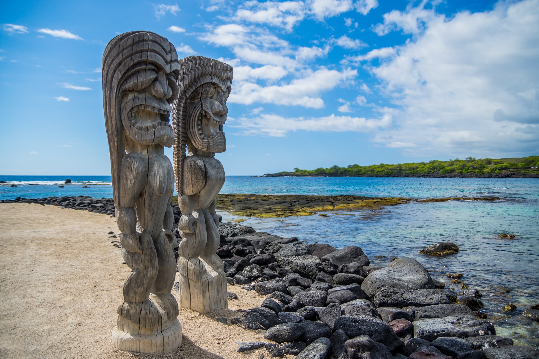 At Puʻuhonua o Hōnaunau National Historical Park, two carved wooden ki‘i stand next to the water to represent gods, deities, and ancestors.