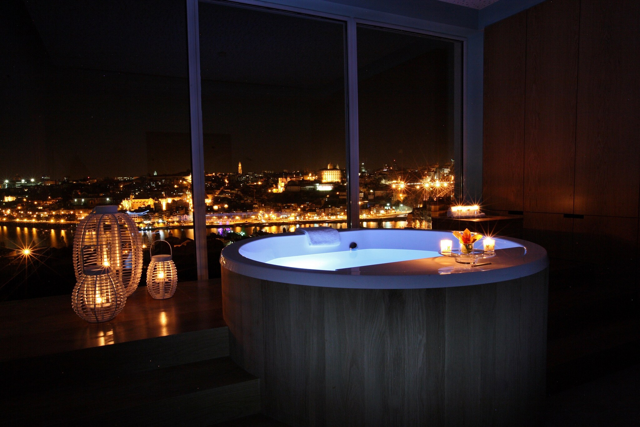 In the dark barrel bath treatment room, candles line the room and the large hot tub overlooks floor-to-ceiling windows showing the city.