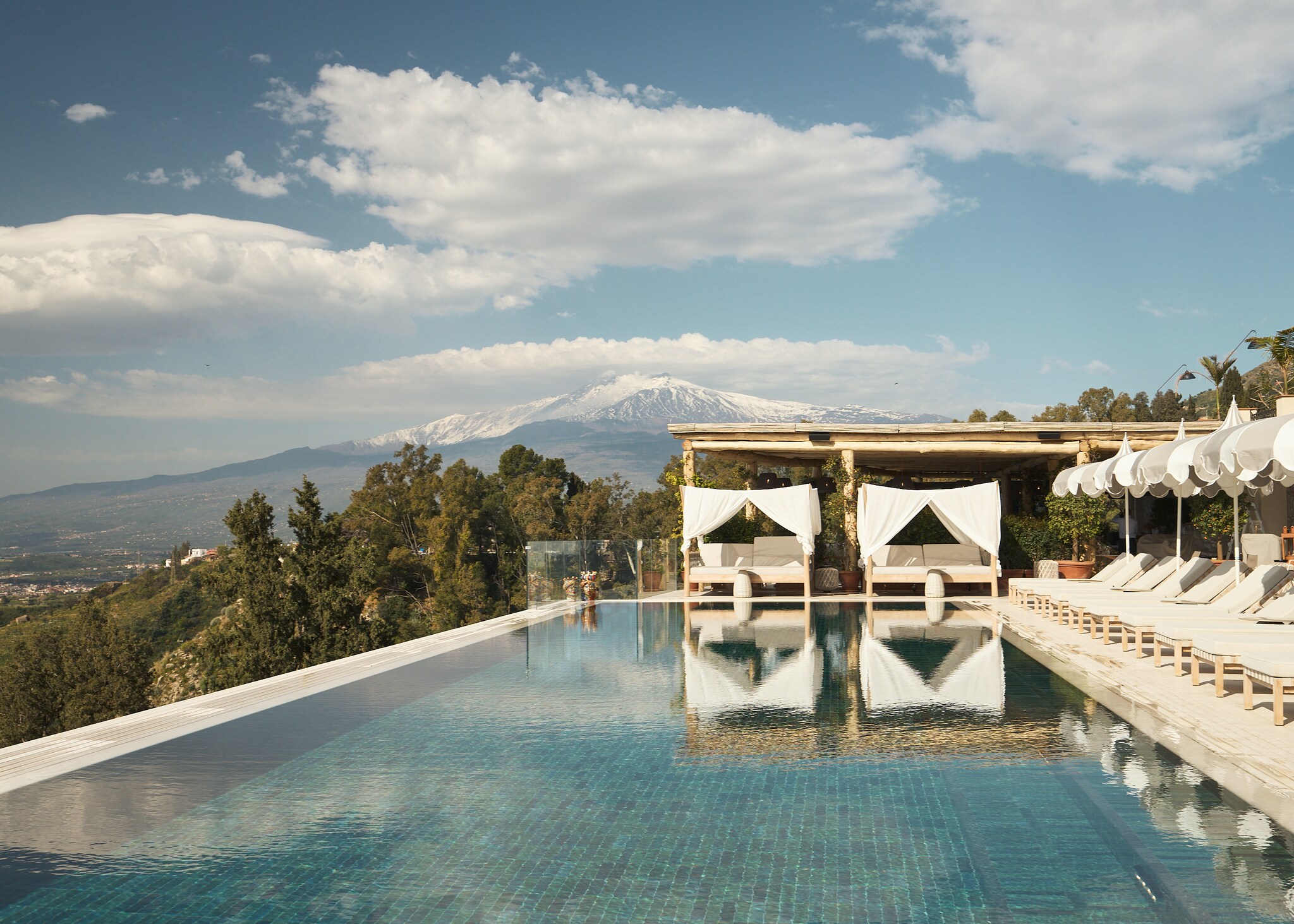 A rectangular infinity pool extends to the edge of a hillside. Sun beds and white umbrellas line one side, with cabanas at the end.