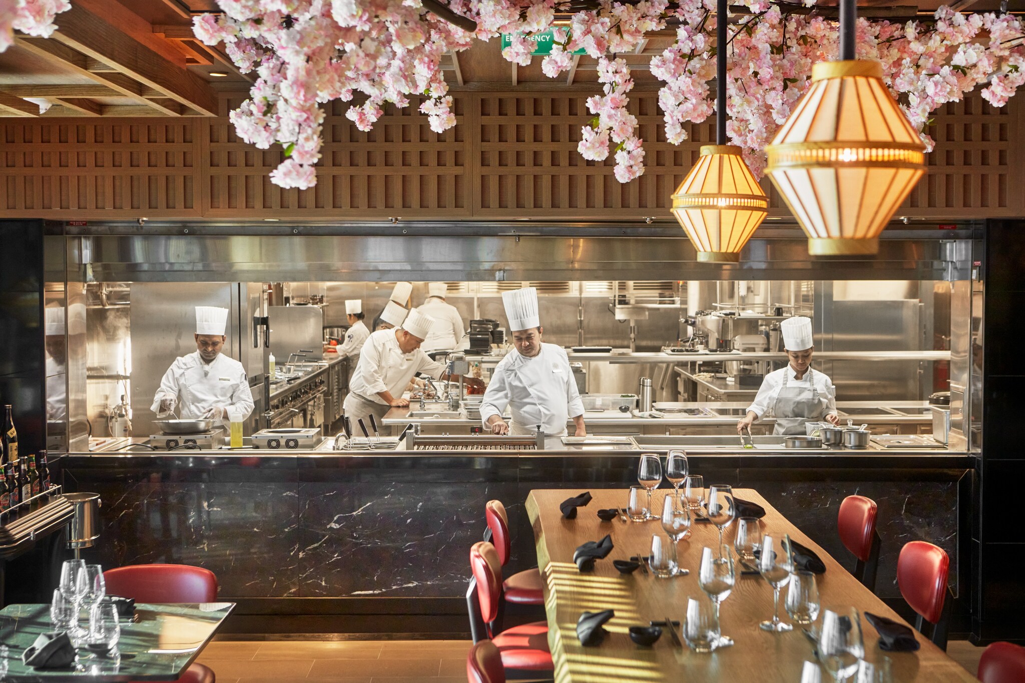 Past an elegant wooden dining table, under a canopy of pink blossoms, a window in the wall reveals a kitchen full of chefs at work.