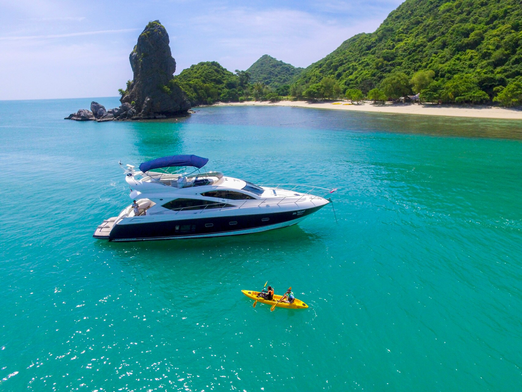 Aerial view of a boat anchored in turquoise waters near a sandy beach. Two people in a yellow kayak paddle towards it.